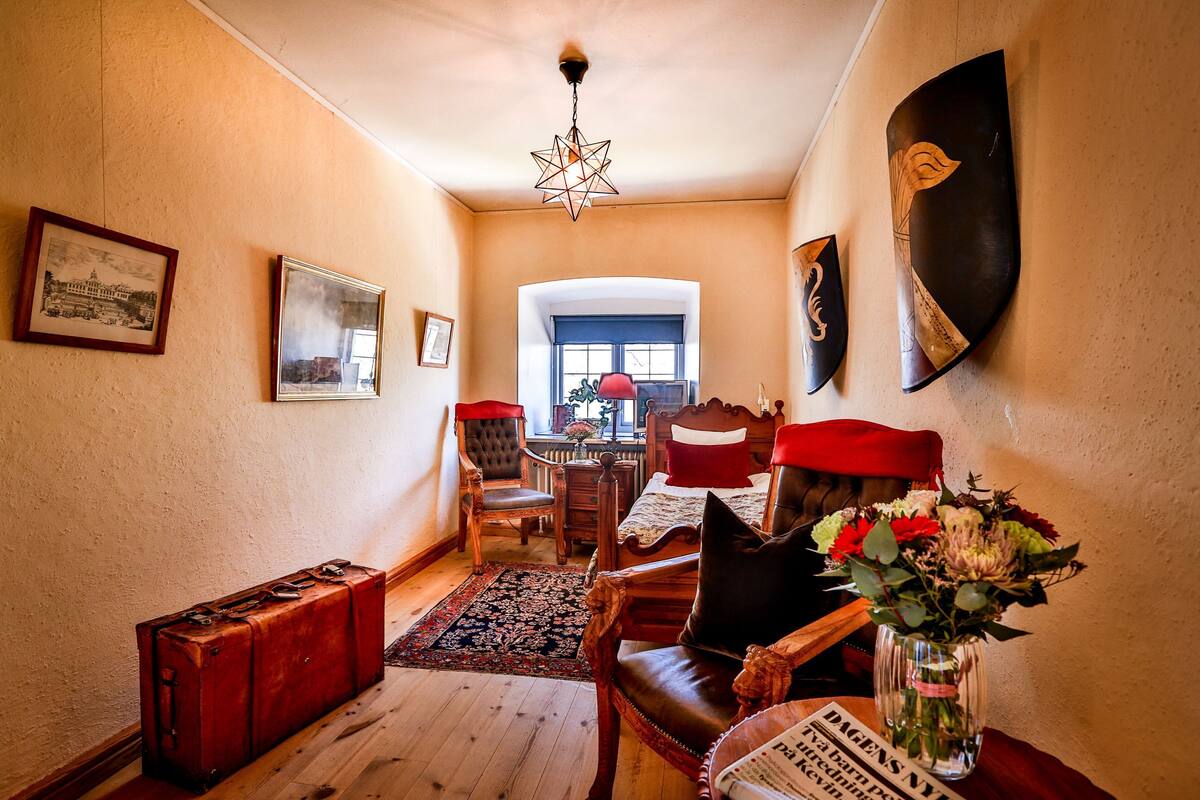 Ein Zimmer mit Holzboden, einem roten Koffer, einem Holzstuhl, einem Bett mit roter Kopfteil, einem Tisch mit Blumenvase und einem gerahmten Bild an der Wand.