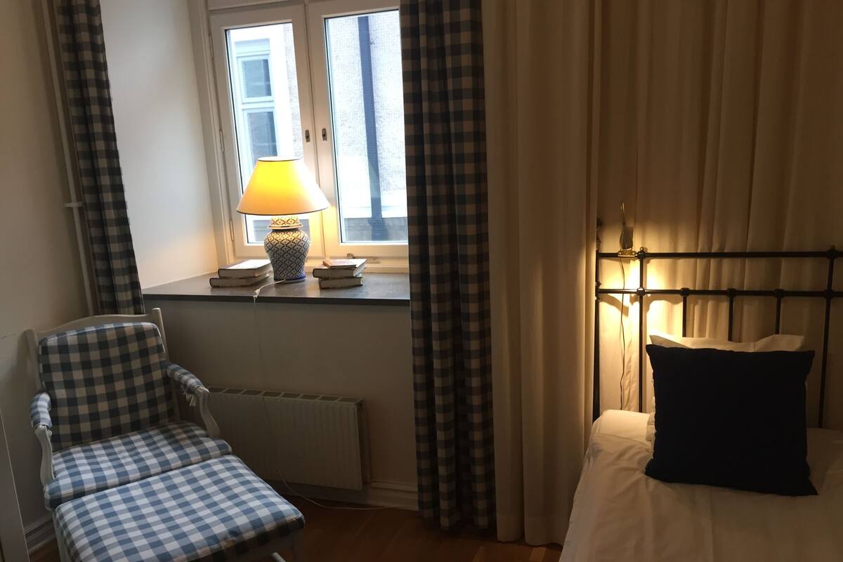 Ein Hotelzimmer mit einem Schachbrettmuster-Sessel, einem Bett mit schwarzem Kopfkissen, einem Fenster mit Vorhängen und einer Lampe auf der Fensterbank.