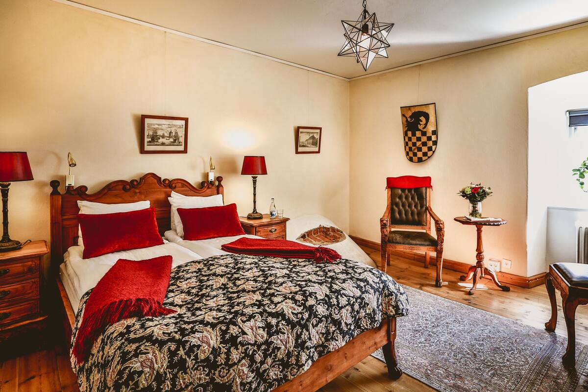 Ein Schlafzimmer mit einem großen Bett, einem Holzkopfstück, zwei Nachttischen mit Lampen, einem Sessel, einem kleinen Tisch mit einer Vase und einem Kleiderständer.