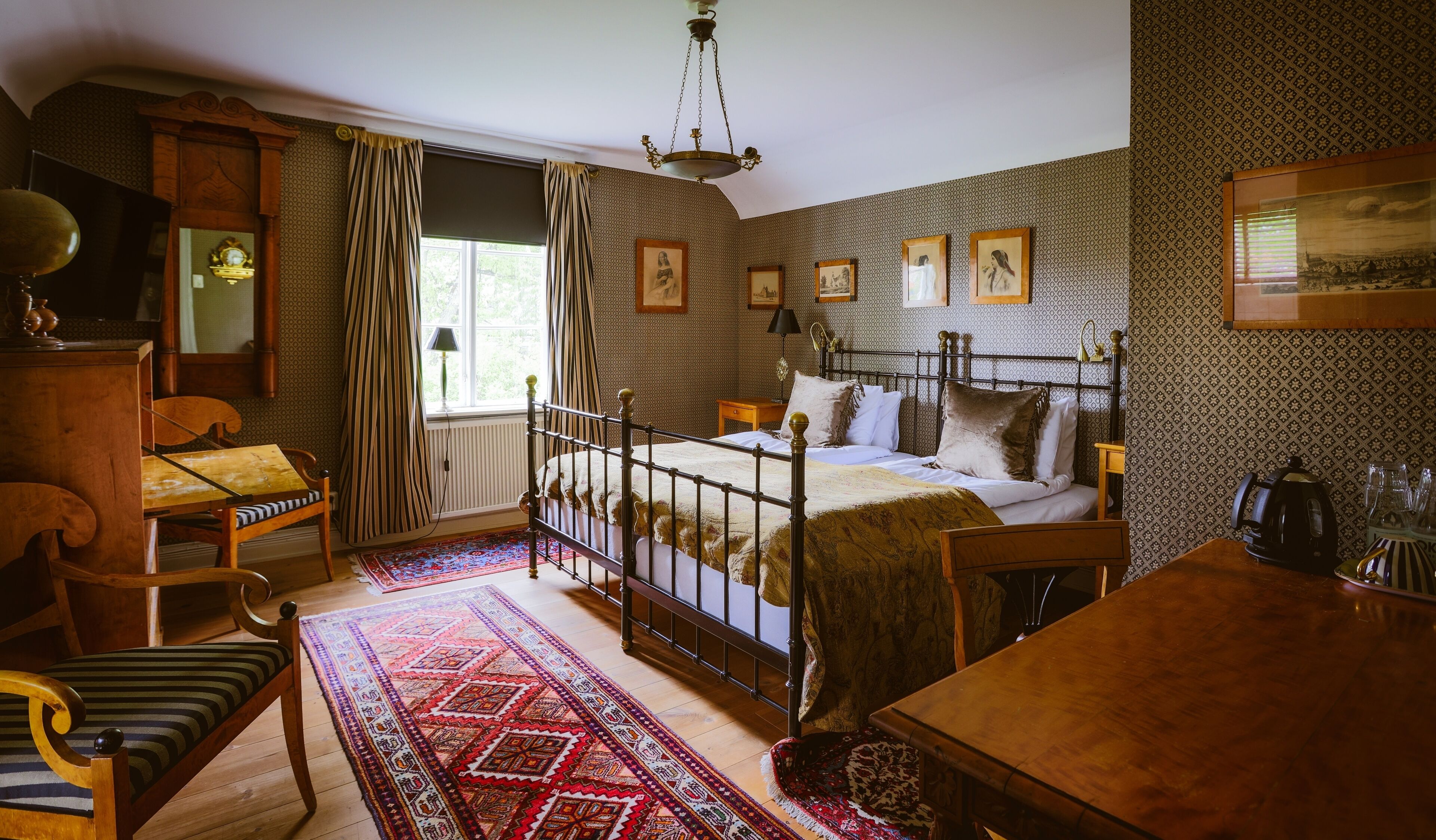 A traditional bedroom with a four-poster bed, a wooden desk, a chair, a patterned rug, and a window with curtains.