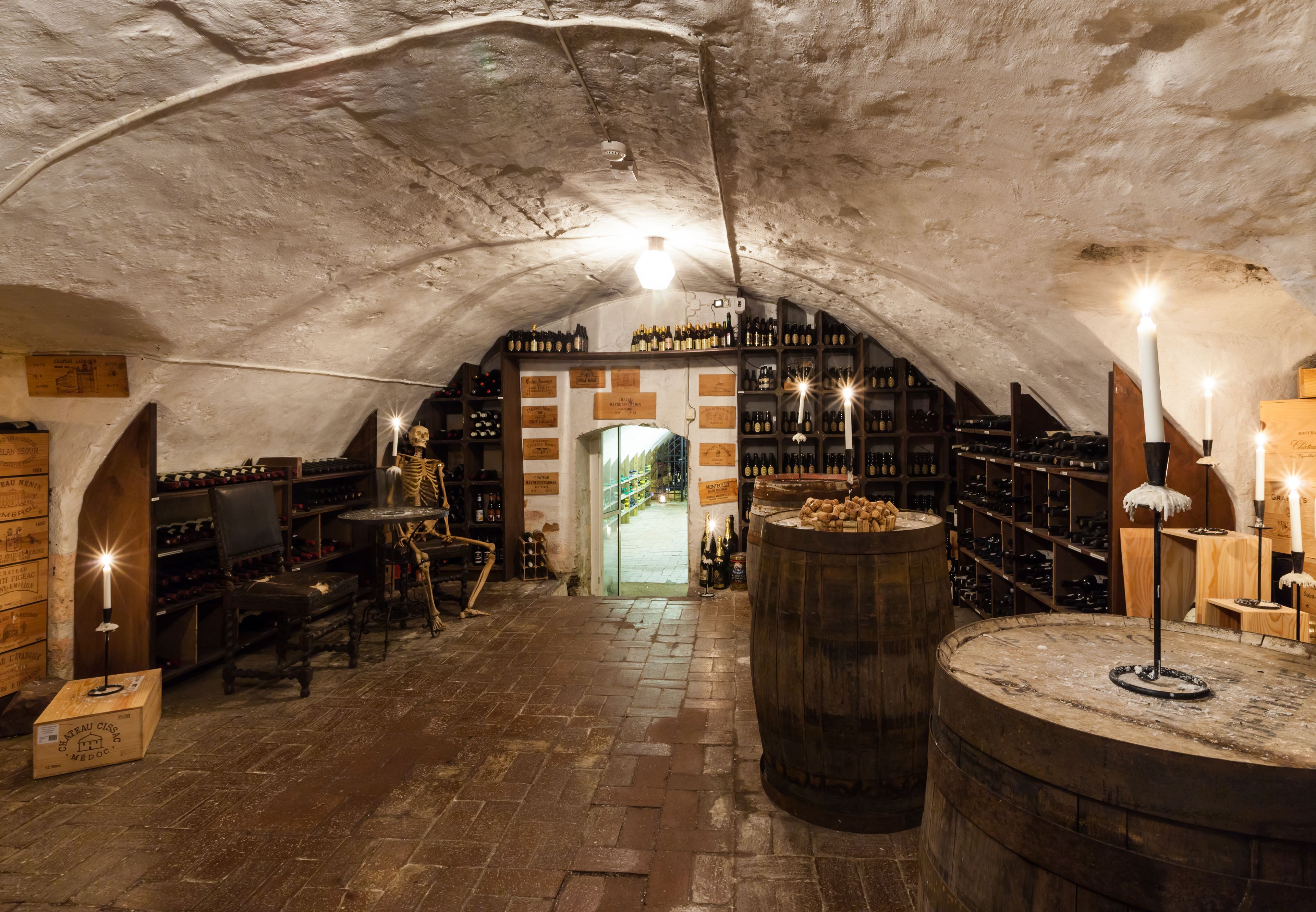 Une cave à vin souterraine, avec des barriques en bois, des étagères remplies de bouteilles de vin et un sol en briques.