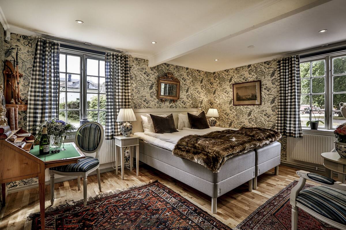 Ein Schlafzimmer mit einem großen Bett, einem Schreibtisch mit Stuhl, einem Fenster mit karierten Vorhängen und einer Wand mit Muster.