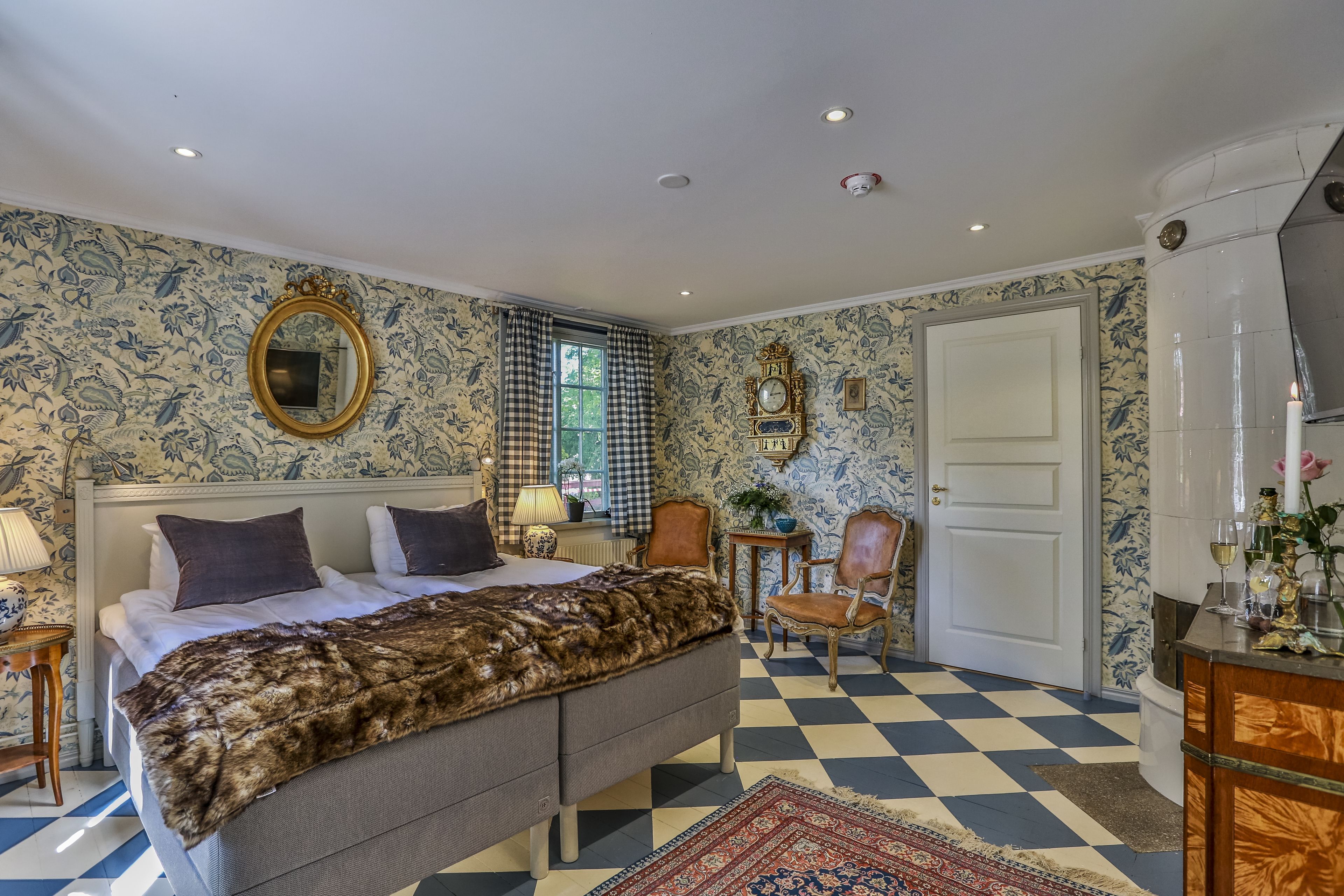 Un dormitorio con piso a cuadros, una cama con un plaid de piel, un espejo, un reloj y una puerta.