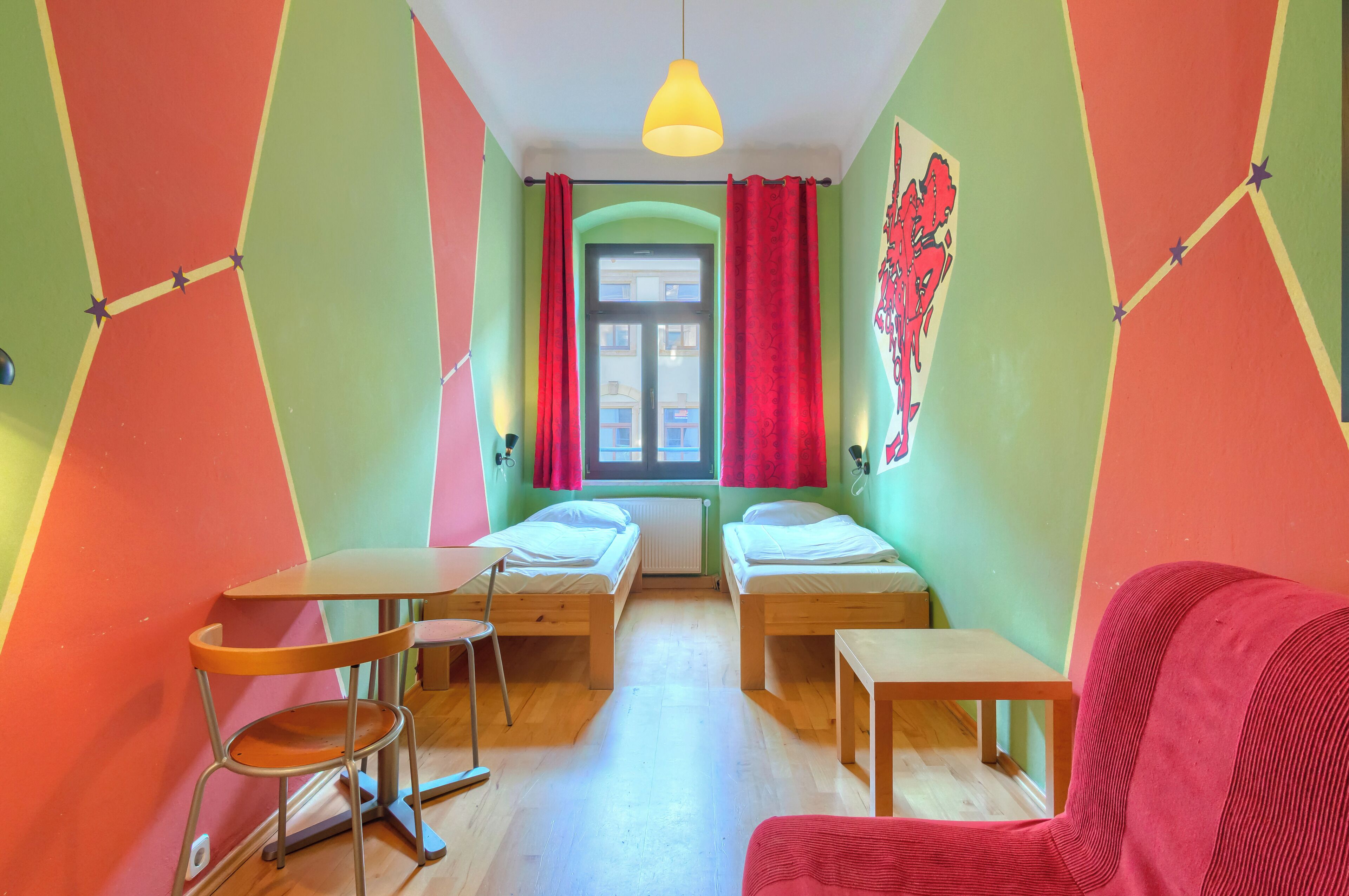 A room with two beds, a small table, a chair, a hanging light fixture, and a window with curtains.