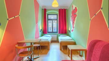 A room with two beds, a small table, a chair, a hanging light fixture, and a window with curtains.