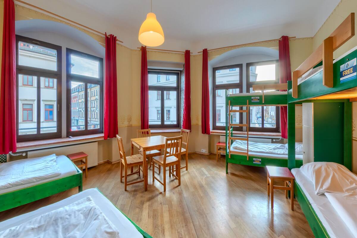 A dormitory room with bunk beds, a dining table with chairs, and large windows with red curtains.