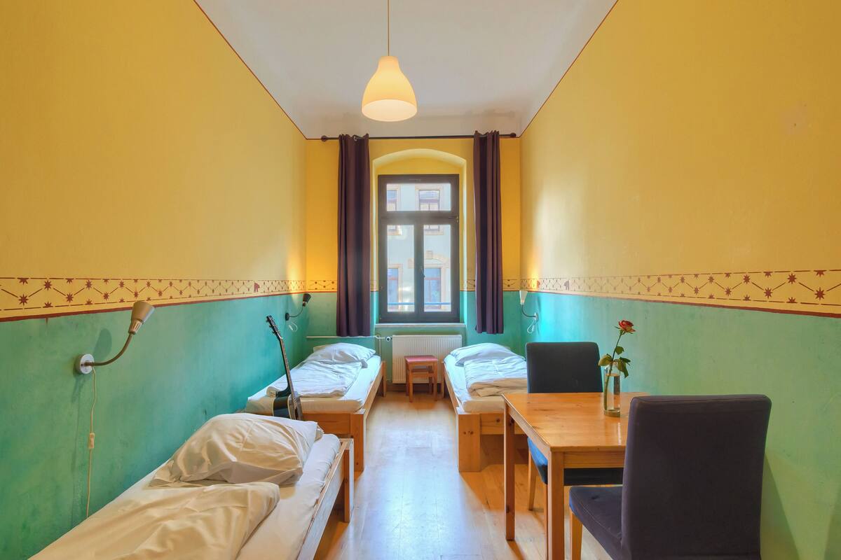A room with two single beds, a wooden table, a chair, a window with curtains, and a hanging light fixture.