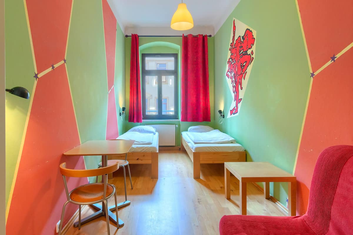 A small, brightly colored room with two single beds, a small table, a chair, and a window with curtains.