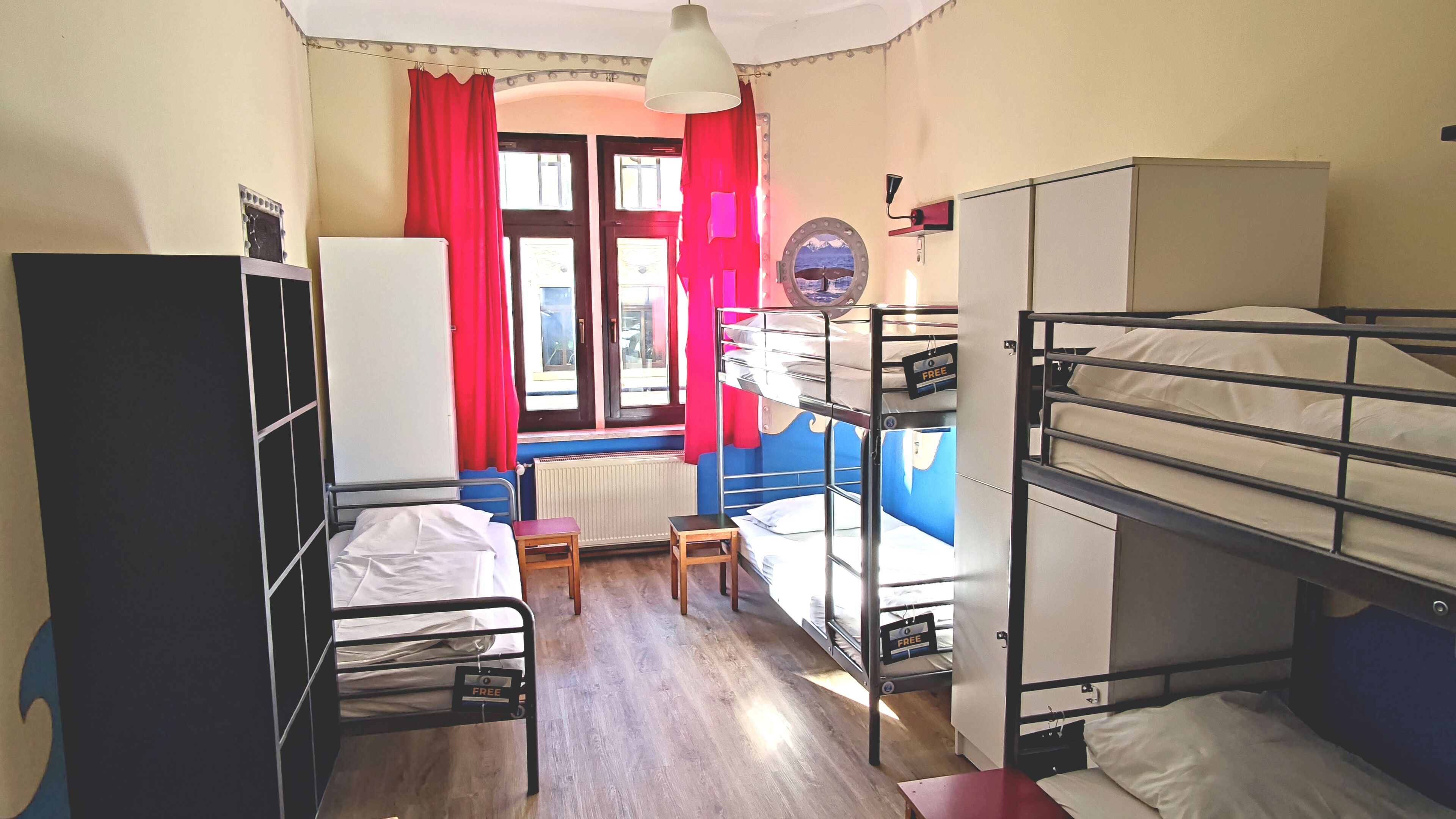 A dormitory room with bunk beds, a desk, a chair, a radiator, and a window with red curtains.