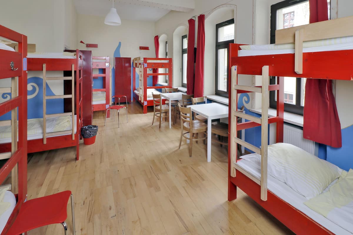 A dormitory room with bunk beds, a desk, chairs, and a window with curtains.