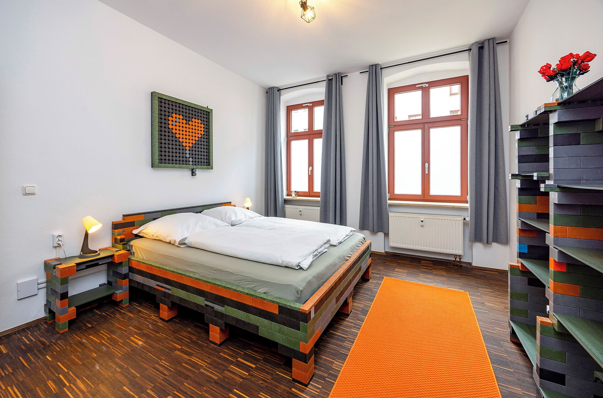 Un dormitorio con cama, mesita de noche, una obra de arte en la pared, ventana con cortinas y una estantería.