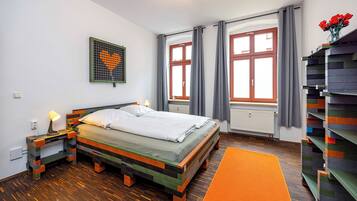 Un dormitorio con cama, mesita de noche, una obra de arte en la pared, ventana con cortinas y una estantería.