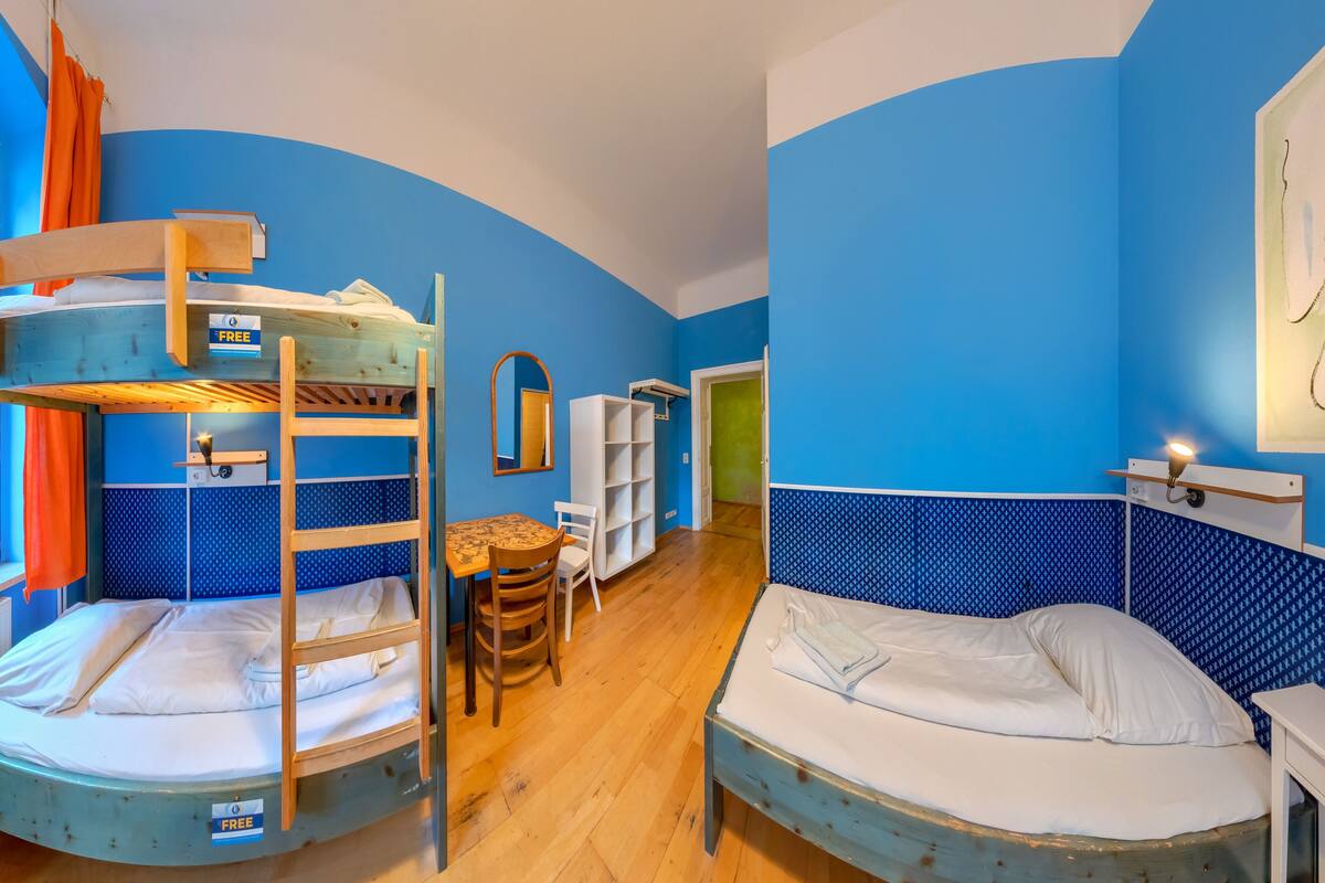 A bunk bed room with a wooden ladder, a small table, and a chair. There is a window with orange curtains and a view of buildings outside.