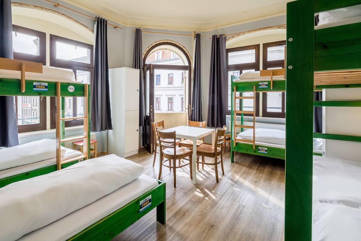 A dormitory room with bunk beds, a dining table, chairs, and a view of the city through large windows.