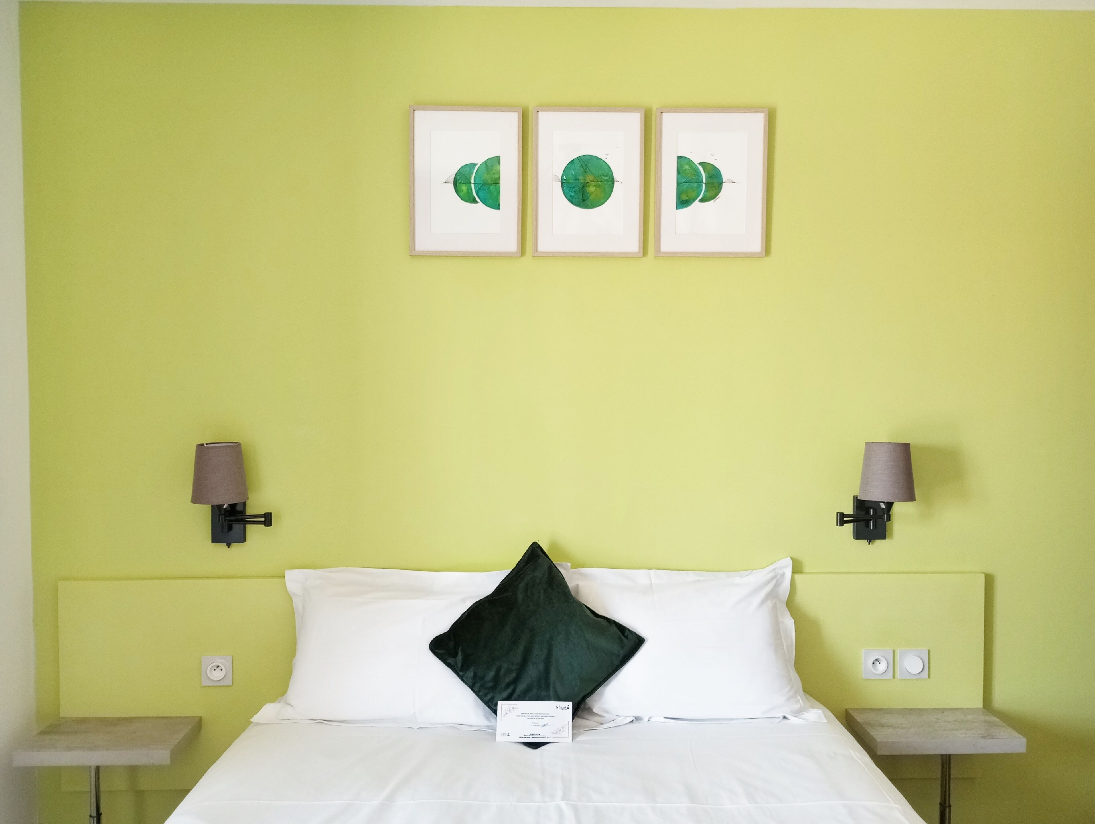 Quarto de hotel com uma cama, duas mesas de cabeceira, um criado-mudo, uma parede verde e três quadros emoldurados.