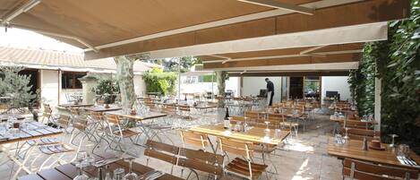 An outdoor dining area with multiple tables and benches, some covered by umbrellas, and a person walking in the background.