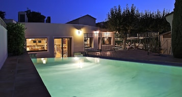 Una piscina con zona de patio, un área de estar al aire libre cubierta y un edificio con varias puertas y ventanas.