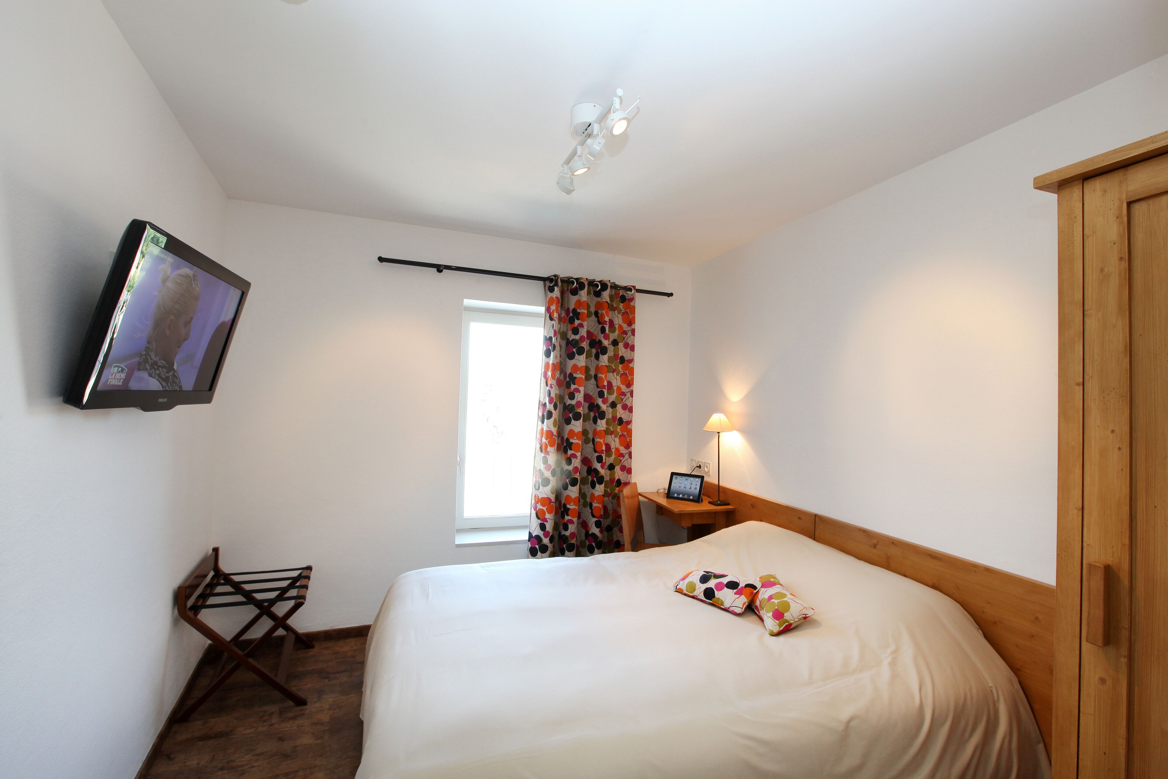 Quarto de hotel com cama, televisão na parede, armário de madeira, mesinha com abajur e janela com cortinas.