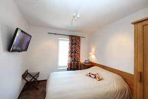 Habitación de hotel con cama, televisión montada en la pared, armario de madera, mesita con lámpara y ventana con cortinas.