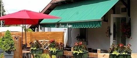 An outdoor seating area with wooden tables and chairs, green awnings, and potted plants.