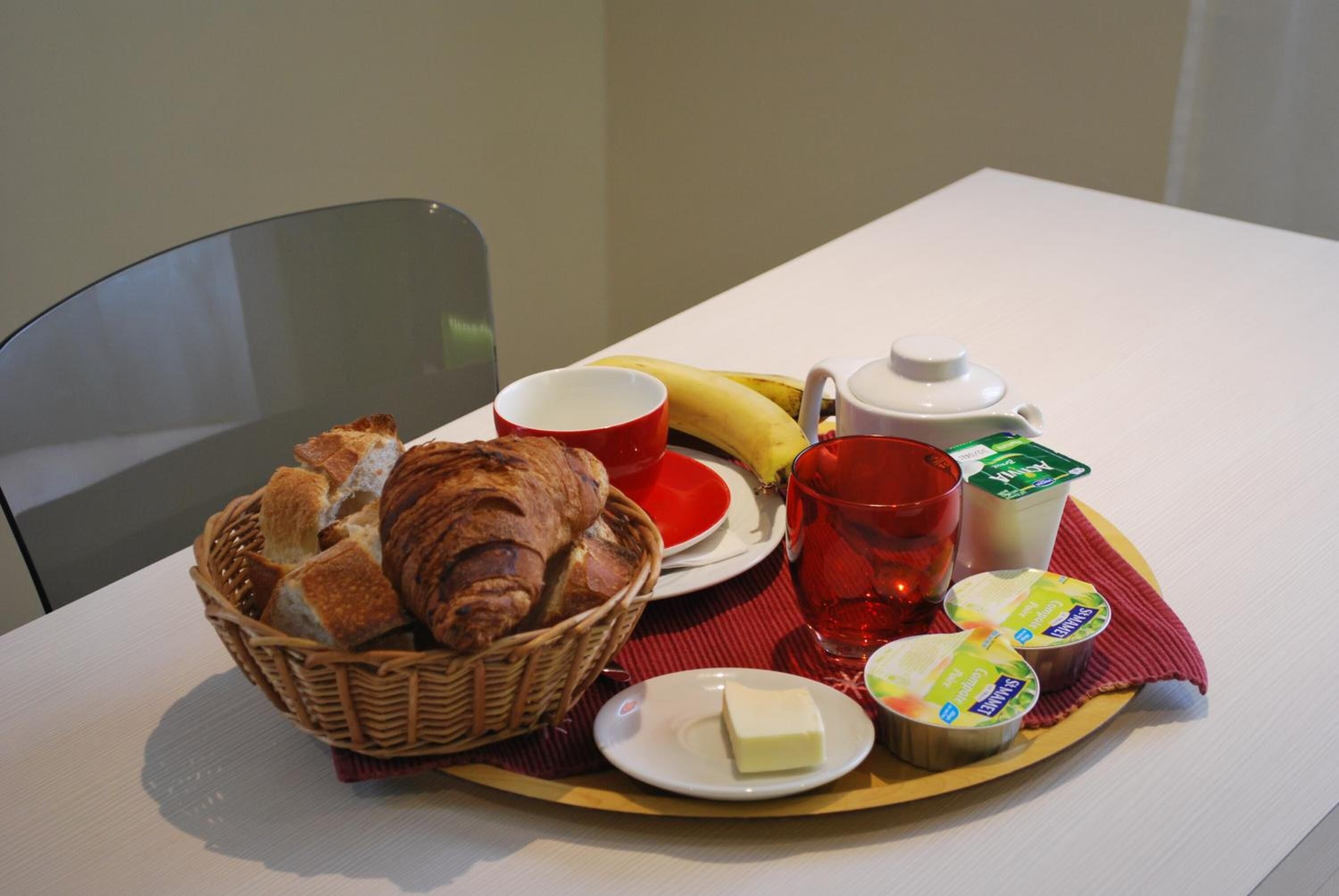 En bakke med morgenmad, bestående af brød, smør, yoghurt, en tepotte, en kop og en banan.