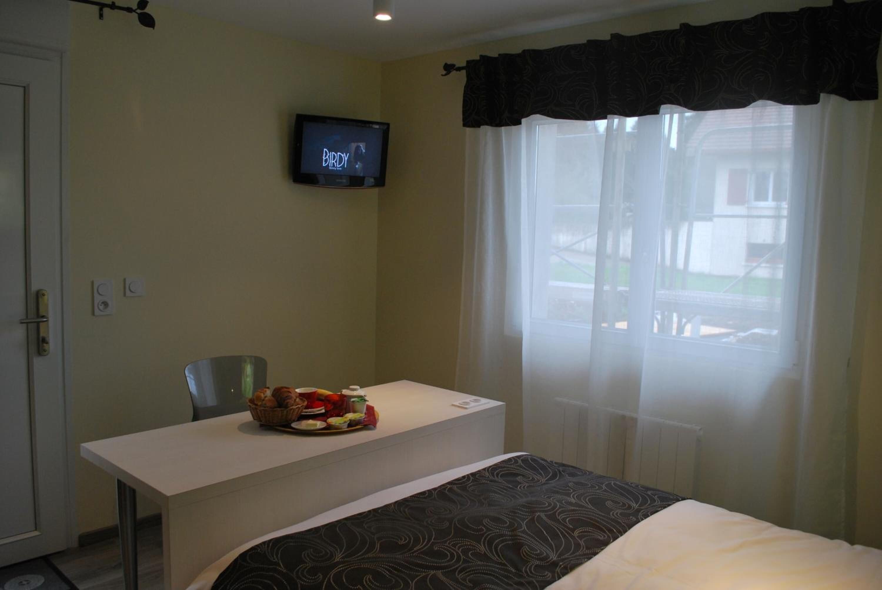 Una habitació d'hotel amb un llit, una taula de nit amb articles per esmorzar, un televisor a la paret i una finestra amb cortines translúcides.