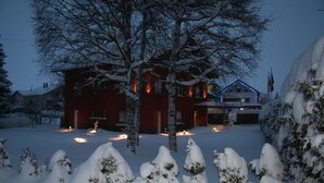 A snow-covered landscape with a red building featuring a blue-lit entrance and a visible flag. - Hôtel Restaurant Au Perce Neige (Bonnetage)