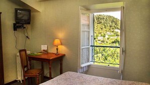A hotel room with a bed, a desk with a lamp, a chair, a TV mounted on the wall, and a window with a view of a green landscape. - Le Pont d'Or (Barjols)