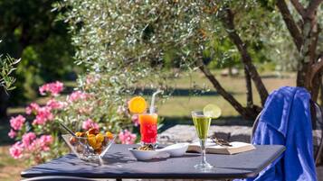 Una mesa al aire libre con un bol de fruta, un vaso de bebida roja con una rodaja de naranja, y un vaso de bebida amarilla con una rodaja de lima.