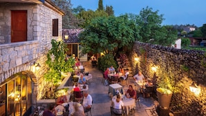 Un área de comedor al aire libre con muros de piedra y mesas, rodeada de vegetación y un edificio de piedra.