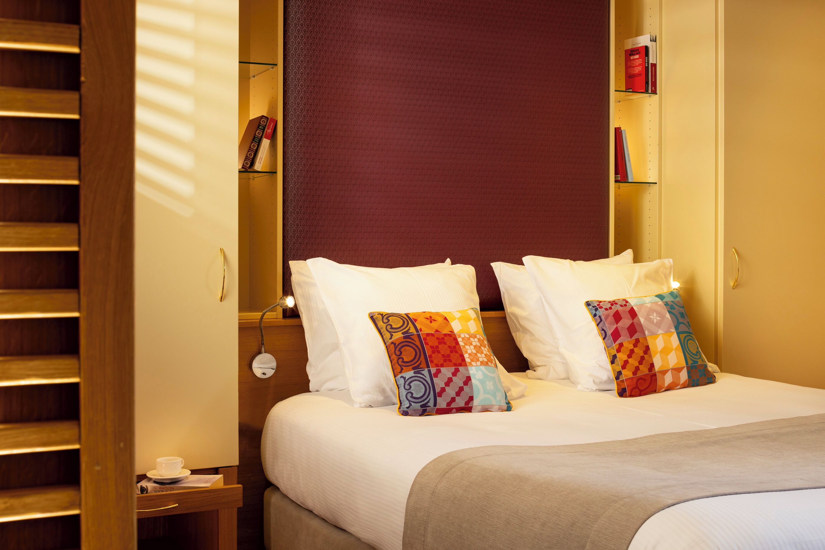A neatly made bed with white linens and colorful patterned pillows, a bedside table with a cup and a book, and a bookshelf with books and a decorative item.