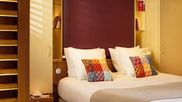 A neatly made bed with white linens and colorful patterned pillows, a bedside table with a cup and a book, and a bookshelf with books and a decorative item.