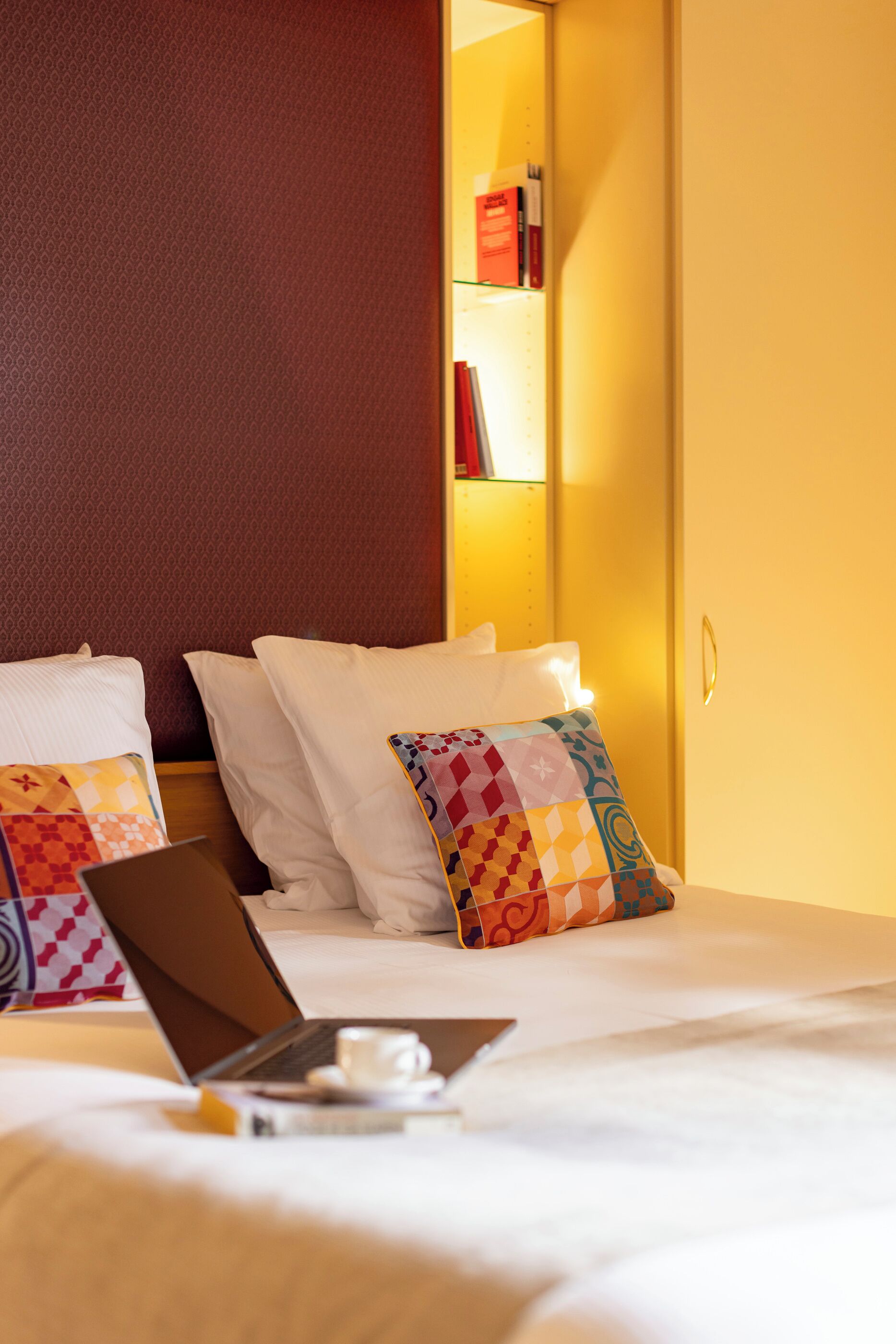 A hotel room with a bed, pillows, a laptop, and a cup on the bedside table.