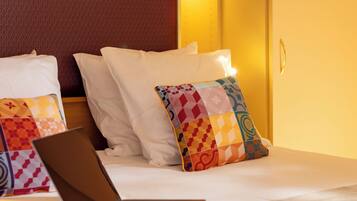 A hotel room with a bed, pillows, a laptop, and a cup on the bedside table.