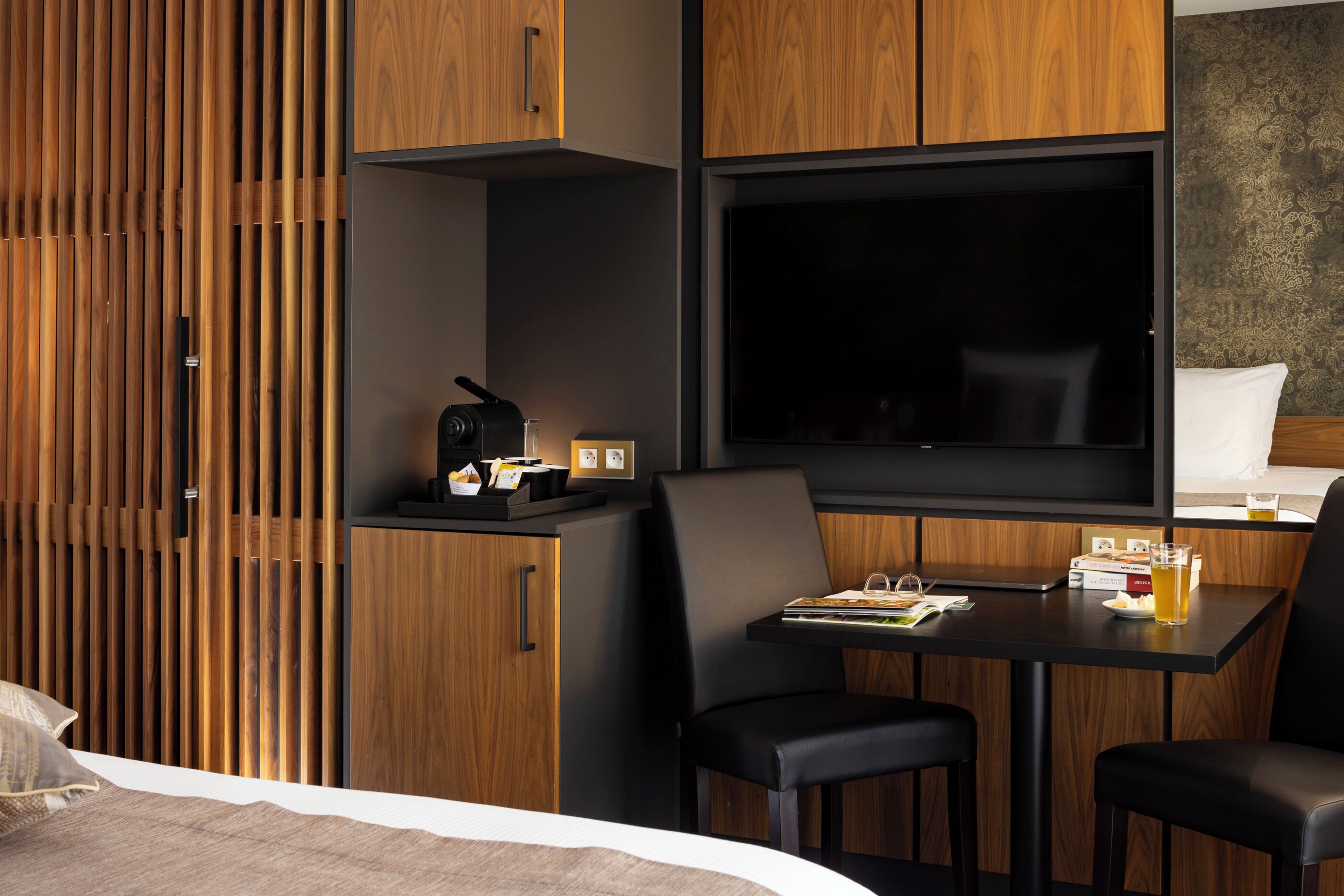 A modern hotel room with a wooden slat wall, a flat-screen TV, a small table with a chair, and a bed with a white comforter.