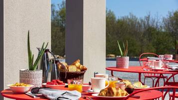Frühstücksaufstellung mit Croissants, Erdbeeren, Orangensaft und Kaffee auf roten Tischen im Freien.