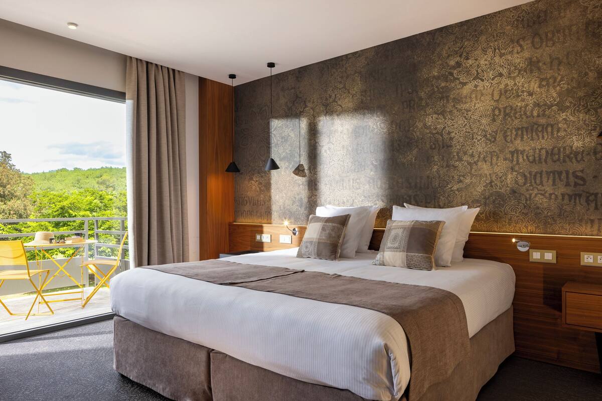 A modern hotel room with a large bed, a balcony with yellow chairs, and a view of greenery.