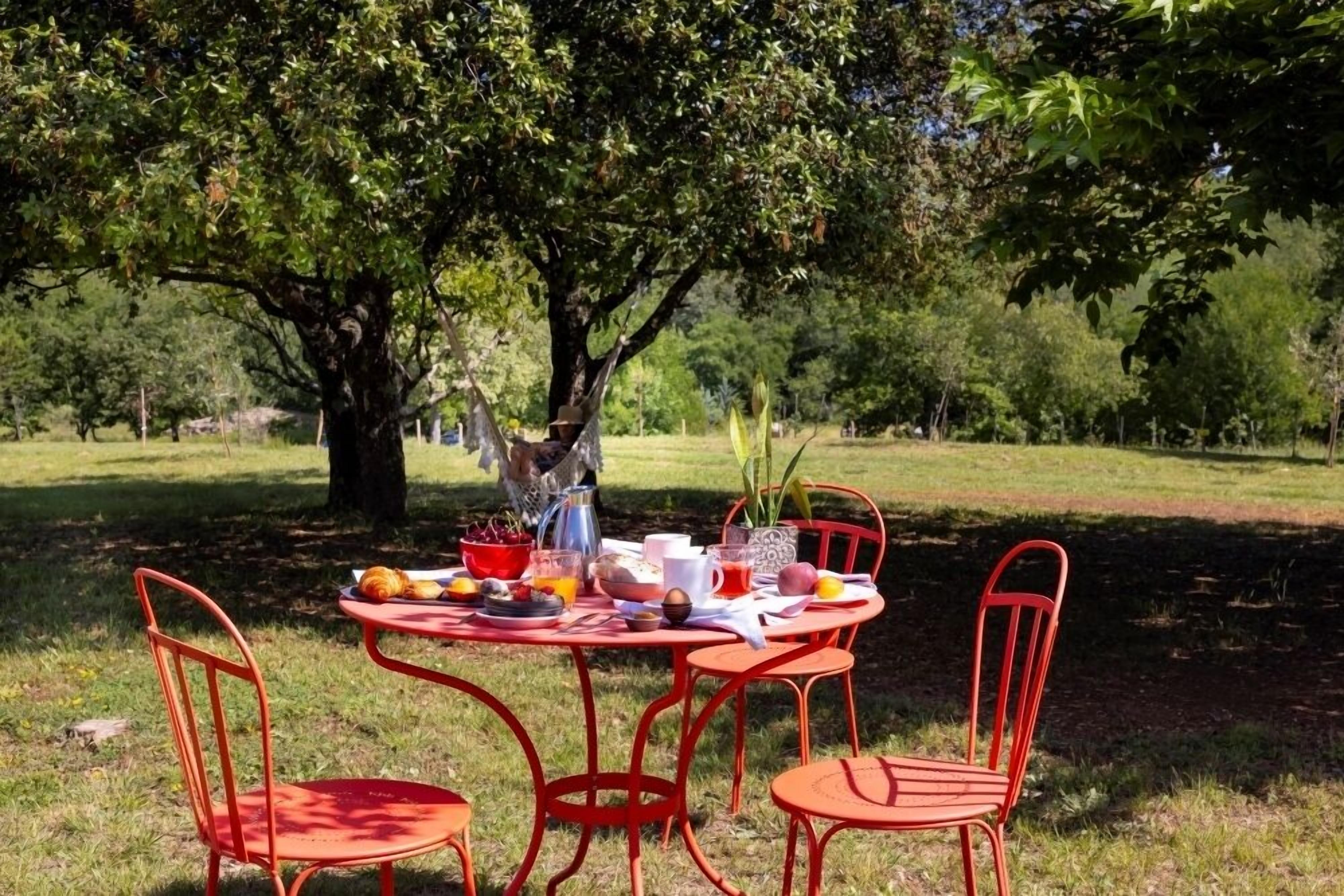 A round table with four red chairs set outdoors under a tree, surrounded by a grassy area.