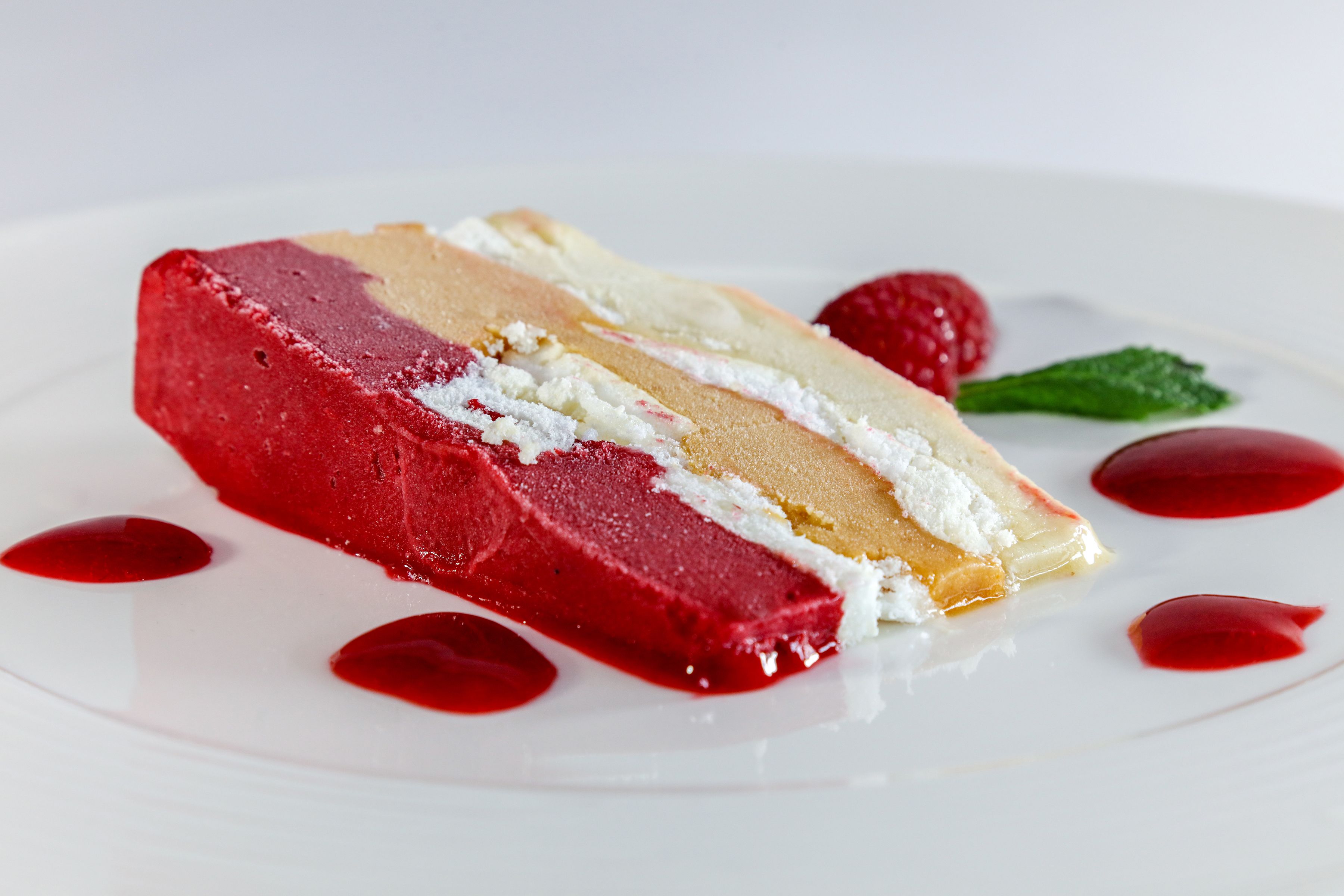 Una porción de pastel en capas con glaseado rojo y blanco, servida en un plato blanco con salsa roja y una hoja de menta.