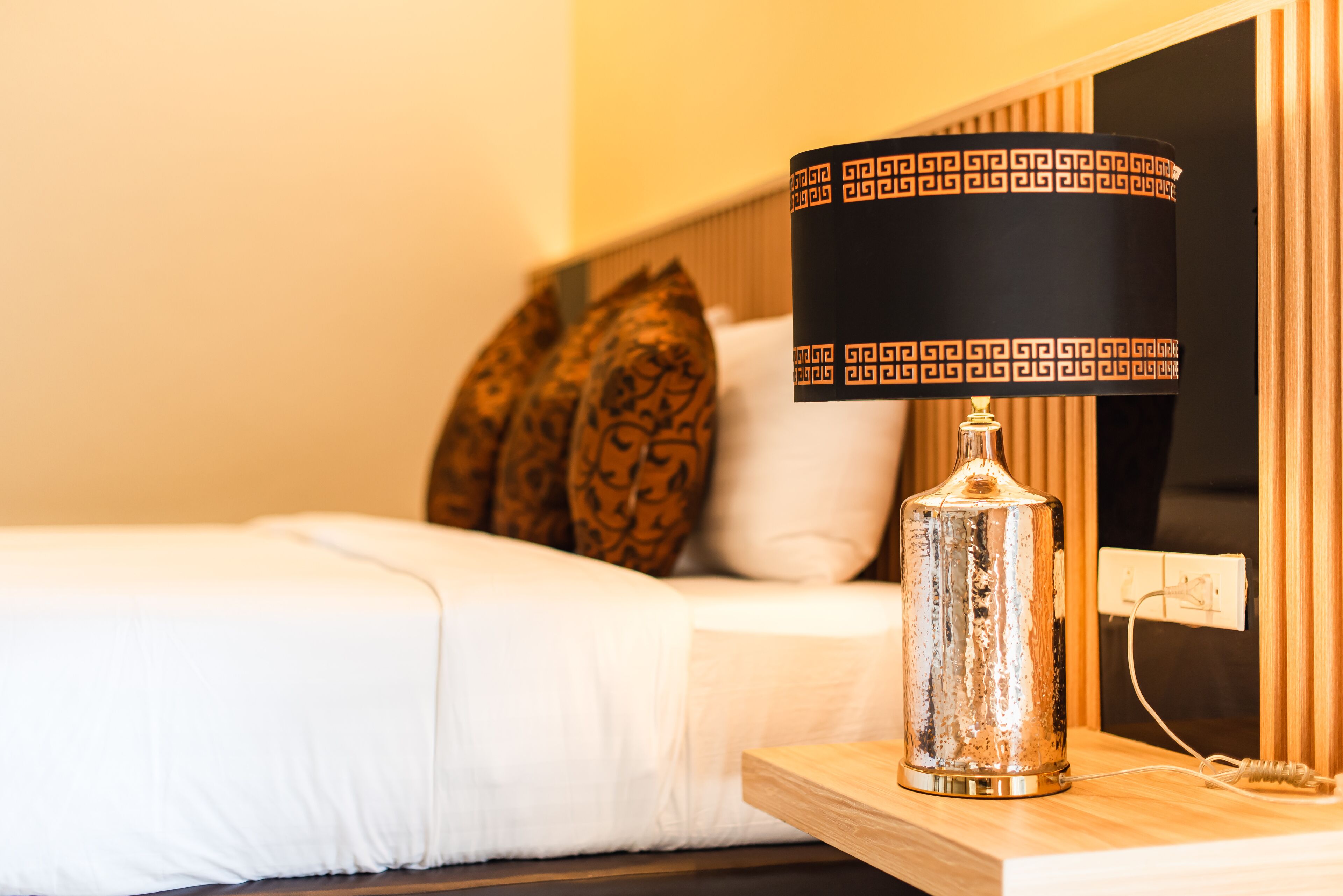A bed with white bedding, a decorative lamp with a patterned shade, and a wooden nightstand.