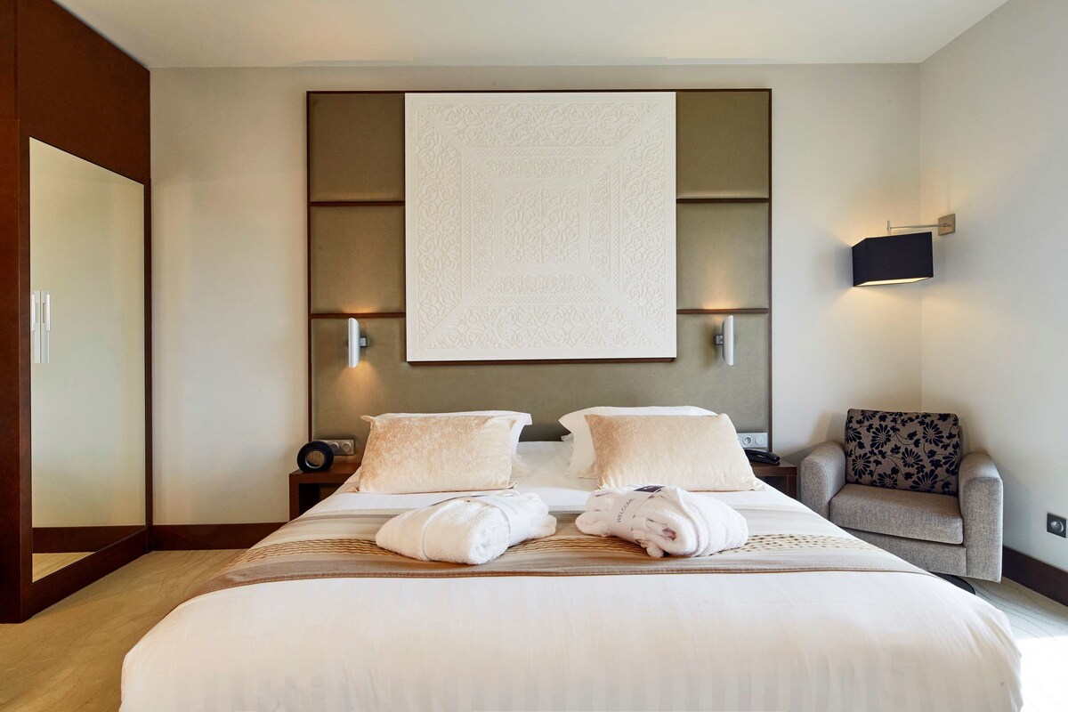 Habitación de hotel con una cama grande, dos mesitas de noche, un televisor montado en la pared y un rincón de descanso con una silla estampada.