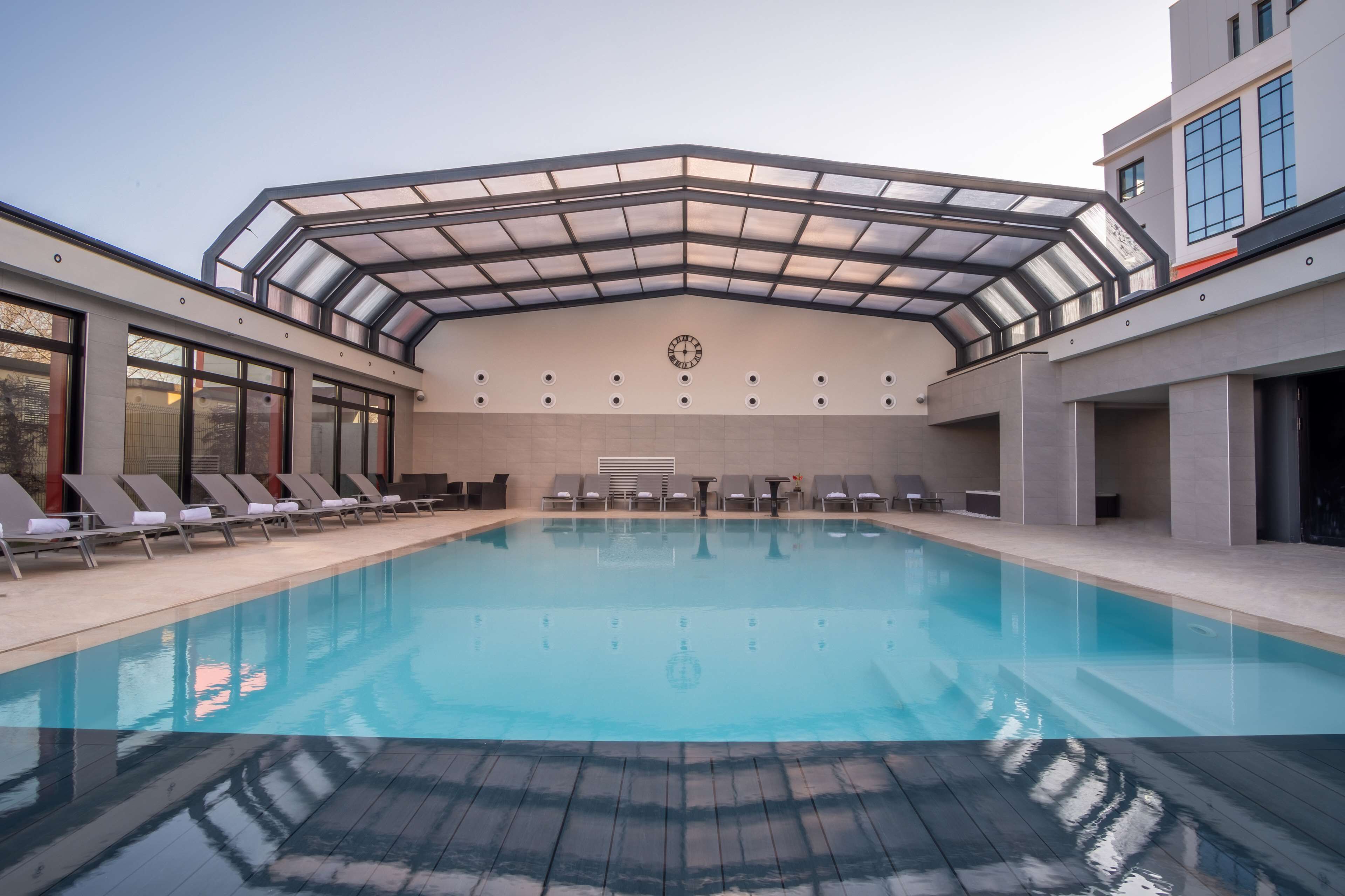 Una piscina interior moderna con techo de cristal, sillas de descanso y un reloj en la pared.