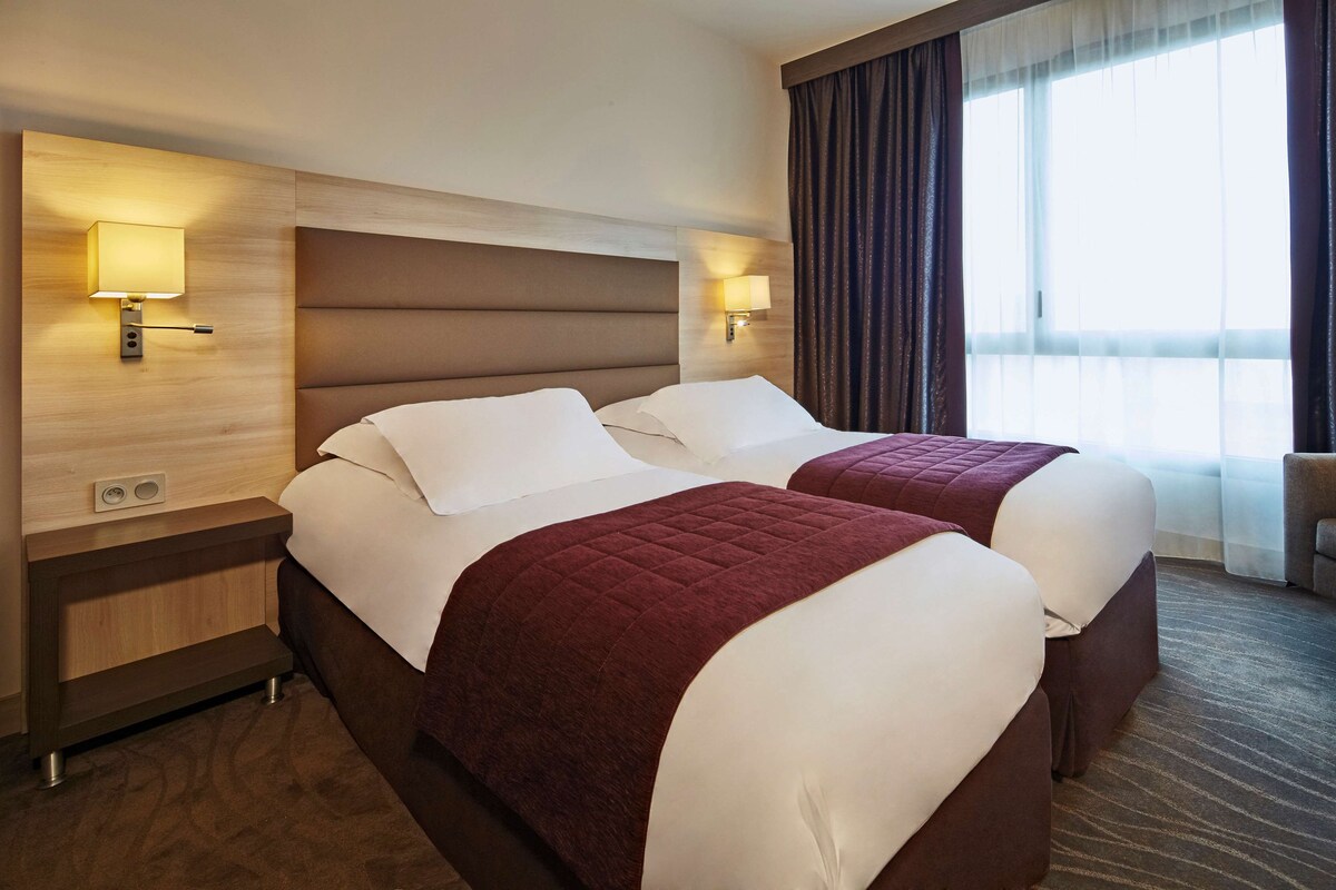 Habitación de hotel con dos camas, cabecero de madera, lámparas fijadas a la pared, un ventanal con cortinas y suelo alfombrado.