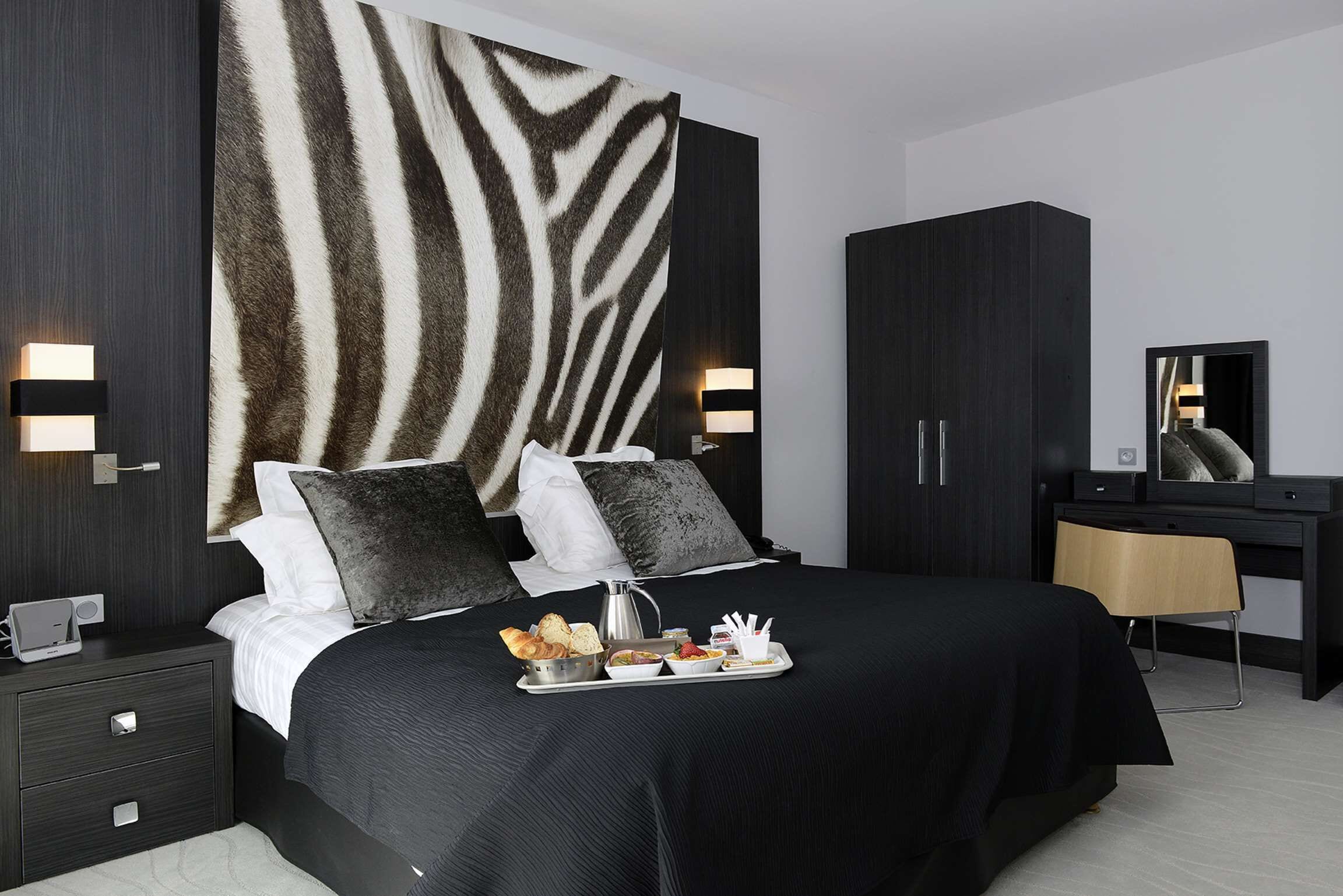 A modern hotel room with a large bed, dark wood furniture, a zebra-patterned wall, and a tray with breakfast items.