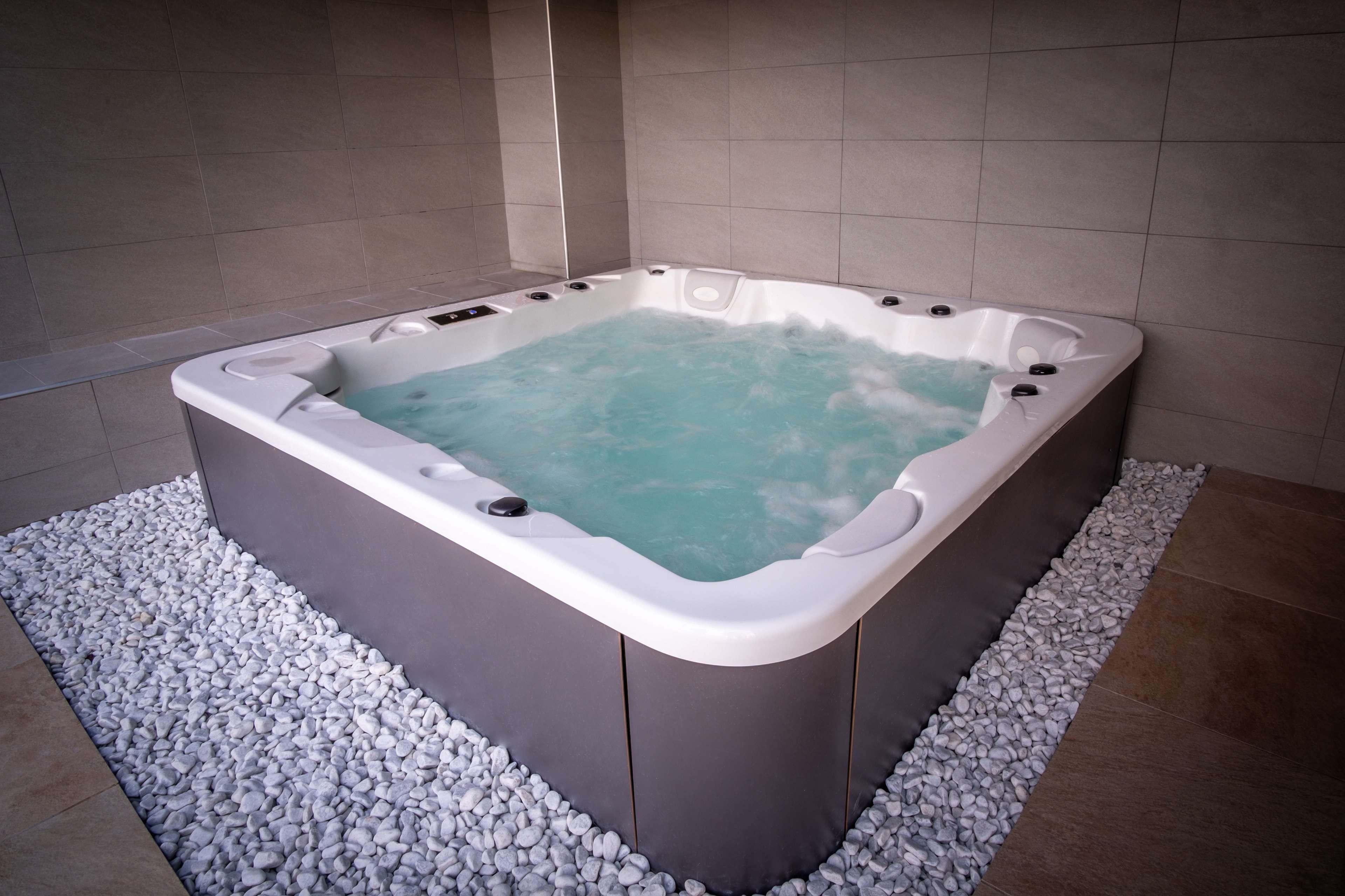 Banyera hidromassatge rectangular amb interior de blau clar, envoltada de petits còdols blancs, situada davant d'una paret de rajola.