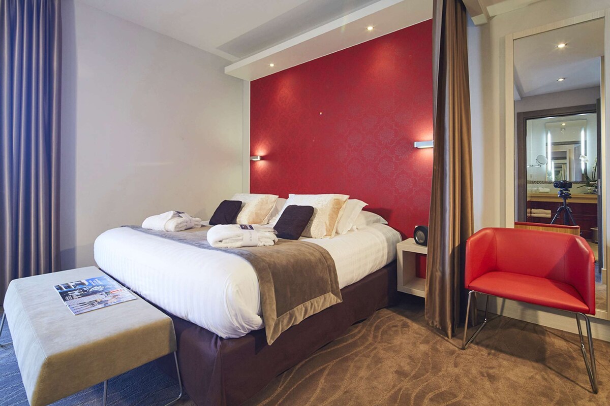 Habitación de hotel con una cama grande, una pared de acento roja, una silla roja y una mesita con una revista.