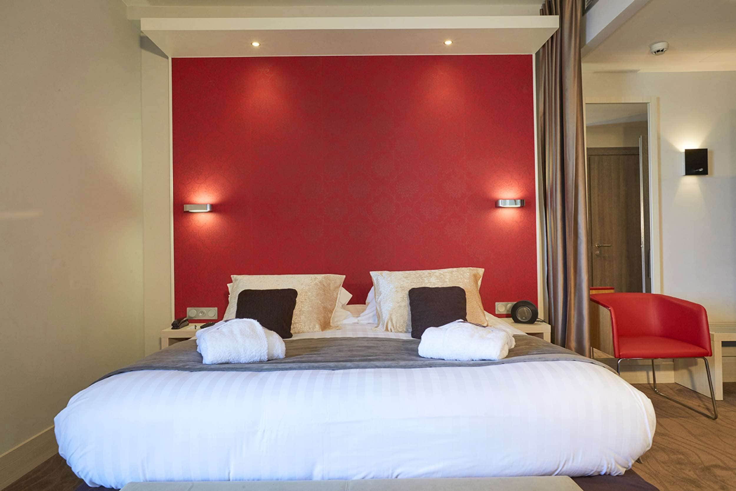 A hotel room with a large bed, red accent wall, and a red chair.
