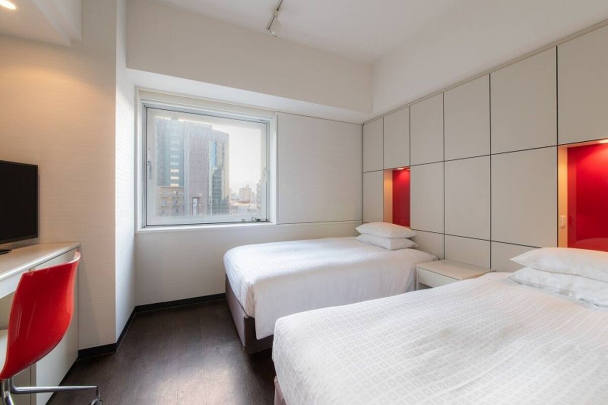 A hotel room with two beds, a desk with a chair, a large window, and a city view.