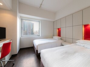 Habitación de hotel con dos camas, un escritorio con una silla, un ventanal grande y vistas a la ciudad.