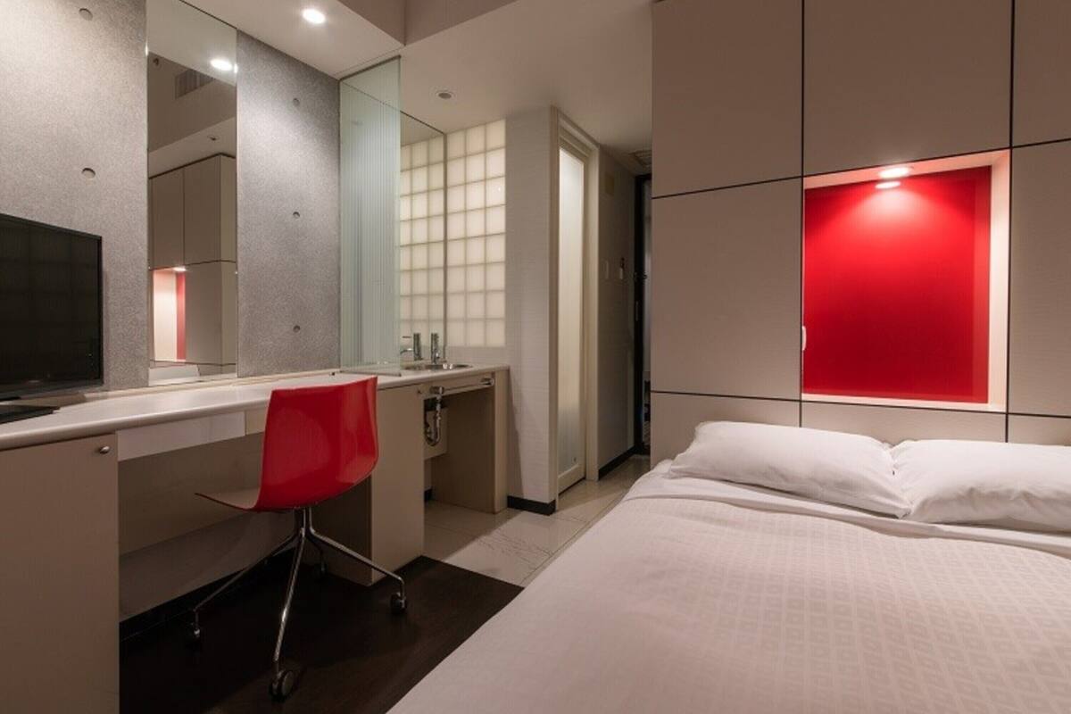 A hotel room with a bed, a desk, a chair, a TV, and a red accent wall.